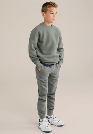 Groene sweatshirt met getextureerde geometrische patronen, gecombineerd met bijpassende joggers, beide met elastische cuffs. Model draagt witte sneakers.
