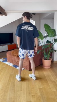 Jūras flotes t-korte ar tekstu "SPEED", baltas šortus ar zilām apļveida rakstām, un pelēkas kedas. iekštelpu vide ar augiem un mēbelēm.