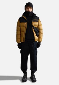 Giubbotto imbottito oro e nero con chiusura a zip, maglione nero sotto, pantaloni cargo, guanti e un berretto nero. Le sneaker completano il look.