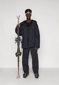 Smučarski komplet s temno modro nepremočljivo jakno, črno osnovno plastjo, črnimi smučarskimi hlačami in črnimi smučarskimi čevlji. Drži lesene smuči z vezmi.