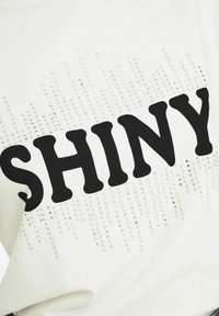 Hvid t-shirt med ordet "SHINY" i fed sort skrift, udsmykket med sølvnitter arrangeret i et gradientmønster.