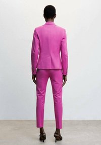 Traje rosa ajustado con americana y pantalones ajustados, diseño a medida, textura suave y botones sutiles, visto desde la espalda.