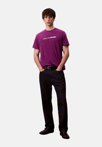 T-shirt en coton violet avec un logo textuel blanc, associé à un jean en denim foncé et des chaussures noires. Le mannequin se tient les mains dans les poches.