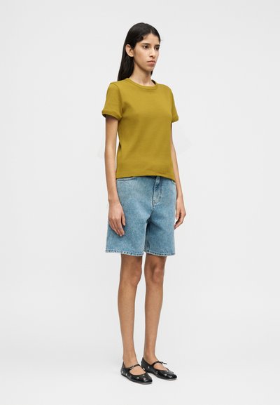 Κίτρινη μπλούζα με κοντό μανίκι και γυρισμένα μανσέτα, συνδυασμένη με ανοιχτόχρωμα τζιν σορτς. Το μοντέλο φοράει μαύρα φλατ παπούτσια με κυκλικές οπές.