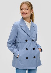 Młoda kobieta uśmiechająca się, ubrana w jasnoniebieską dwurzędową marynarkę z czarnymi guzikami, mająca na sobie białą koszulę i niebieskie dżinsy, z rękami w kieszeniach.