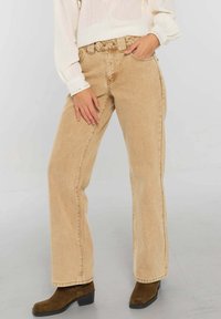 Pantalon en jean beige à jambes larges avec une taille haute, doté de cinq poches et d'un léger effet délavé. Associé à un chemisier blanc texturé à manches longues.