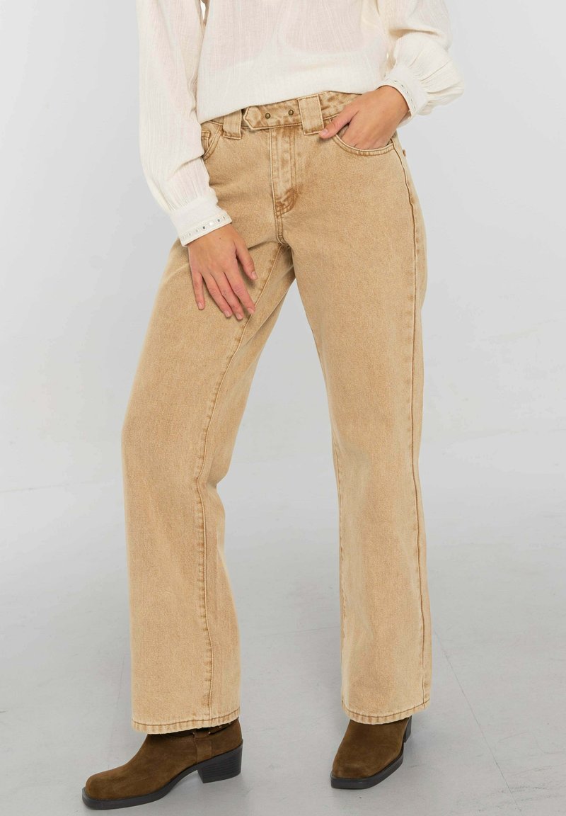 Pantalon en jean beige à jambes larges avec une taille haute, doté de cinq poches et d'un léger effet délavé. Associé à un chemisier blanc texturé à manches longues.