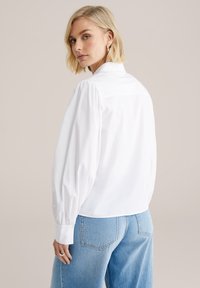 White cotton blouse with puffed sleeves and a back yoke detail, paired with light blue jeans. Smooth texture and classic design.