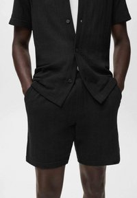Homme portant une chemise bouton-up noire texturée à manches courtes et un short assorti, les mains dans les poches, sur un fond clair uni.
