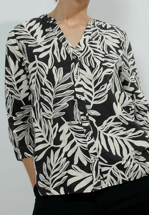 Femme portant une chemise noire boutonnée avec un motif de feuilles blanches épaisses et des manches trois-quarts, debout devant un arrière-plan uni.