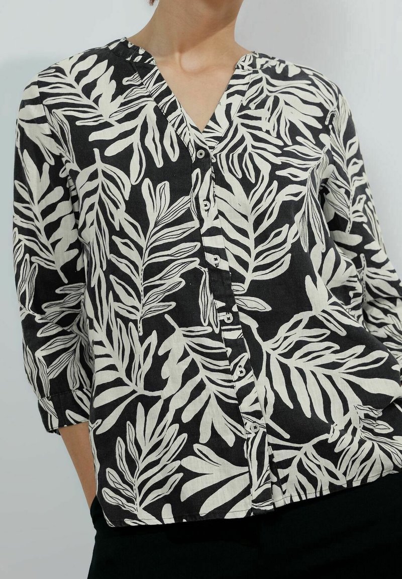 Femme portant une chemise noire boutonnée avec un motif de feuilles blanches épaisses et des manches trois-quarts, debout devant un arrière-plan uni.