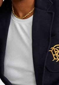 Personne portant une chemise blanche côtelée sous un blazer bleu marine avec des initiales brodées en or et un collier chaîne en or torsadée.