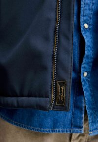 Veste de marine avec une fermeture éclair prononcée, dotée d'un tirette en ton doré et d'une étiquette. En dessous, une chemise en denim bleu avec un design à boutons.