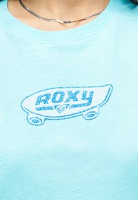 Άτομο που φοράει ανοιχτό μπλε πουκάμισο με μπλε γραφικό skateboard και το κείμενο «ROXY» τυπωμένο στην περιοχή του στήθους.