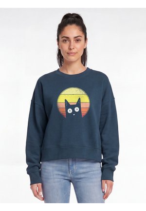Junge Frau mit dunklen Haaren im Pferdeschwanz, die einen dunkelblauen Pullover mit gelbem und orangefarbenem Sonnenuntergang und einer schwarzen Katzen-Grafik trägt, kombiniert mit hellblauen Jeans.