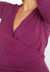 Femme portant un pull cache-cœur à manches longues violet profond, montrée des épaules à la taille avec le bras levé.