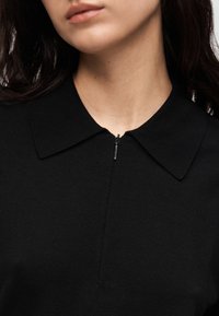 Μαύρο πλεκτό τοπ με γιακά και κοντό φερμουάρ μπροστά. Το ύφασμα φαίνεται λείο, με εφαρμοστή σχεδίαση που αναδεικνύει τη γραμμή του λαιμού.