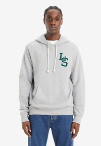 Γκρίζα φούτερ με μπροστινή τσέπη, λευκές κορδόνες και κεντητό πράσινο λογότυπο στη αριστερή πλευρά του στήθους. Φορεμένο με μπλε τζιν.