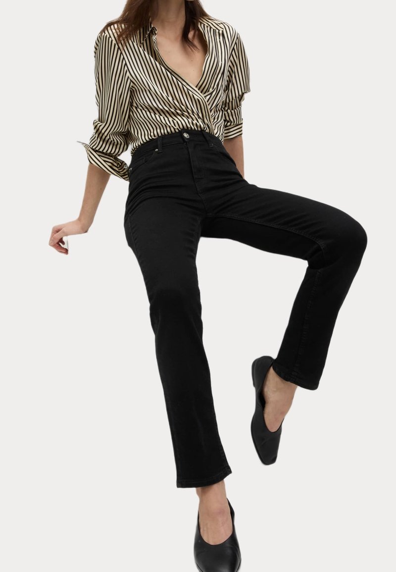 Femme portant un chemisier beige et noir rayé, un jean noir taille haute et des ballerines noires, posant avec une jambe levée devant un fond uni.