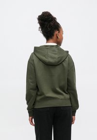 Olívazöld kapucnis pulóver strukturált kivitelben, bordázott mandzsettákkal és szegéllyel, valamint puha tapintású anyagból. Hátul egy finom logó részlet található.