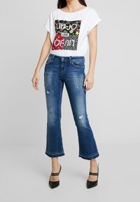 Camiseta blanca con diseño gráfico en negro y rojo. Jeans acampanados azules con lavado desvanecido y detalles desgastados. Zapatos de tacón alto negros.