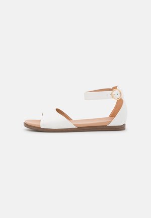 Hvid åbentået sandal med ankelrem, justerbar spænde og tan sål, der har en flad sål og minimalistisk design.