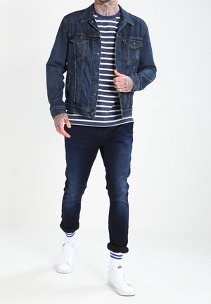 Mann trägt eine dunkelblaue Jeansjacke, ein navyblau gestreiftes Hemd, hochgekrempelte dunkle Jeans, weiße Sneaker und gestreifte Socken und steht vor einem weißen Hintergrund.