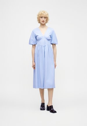 Woman with curly blonde hair wears light blue vertical striped midi dress with puff sleeves and black heeled mules, standing on white background.