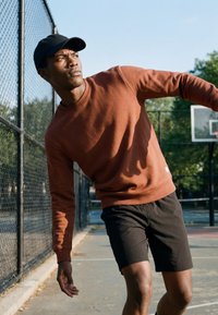 Jeune homme en sweat marron et casquette noire penché de côté sur un terrain de basket en plein air près d'une clôture en grillage.