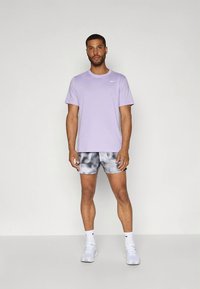 T-shirt desportiva de mangas curtas cor lilás claro, combinada com calções cinzentos com padrão abstracto e ténis brancos. Design simples, ajuste confortável.