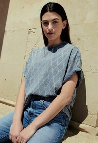 Svetle modrý oversized svetr s diamantovým vzorem, zkombinovaný s modrými džíny s vysokým pasem. Hladký materiál, volný střih.