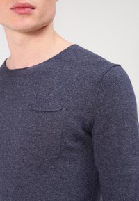 Primo piano di una persona che indossa un maglione a maglia blu scuro con una piccola tasca sul petto e scollo rotondo su uno sfondo semplice.