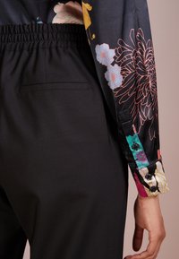 Un pantalon noir à la texture lisse présente une taille froncée. La manche affiche un motif floral coloré sur un fond sombre.