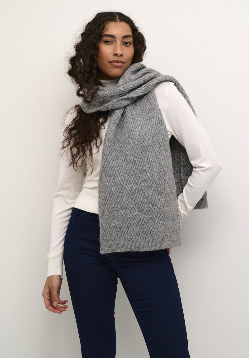 Écharpe en tricot gris arborant un motif en chevrons, texture douce et bords frangés, drapée sur un haut à manches longues blanc.