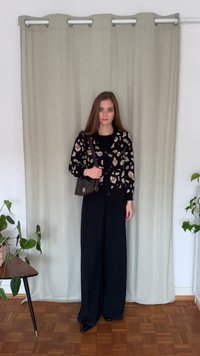 Czarny kardigan z beżowym wzorem liści, czarny top, czarne spodnie o szerokich nogawkach, trzyma ciemną torebkę, stoi na drewnianej podłodze obok rośliny.