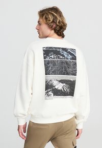 Felpa bianca con una grafica in bianco e nero sul retro, che presenta la topografia di una montagna e fotografie. Vestibilità rilassata con polsini a costine.