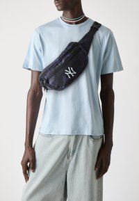 Sac banane noir et camo marine avec un logo blanc proéminent, doté d'une fermeture éclair et d'une bandoulière ajustable, porté sur un t-shirt bleu clair.