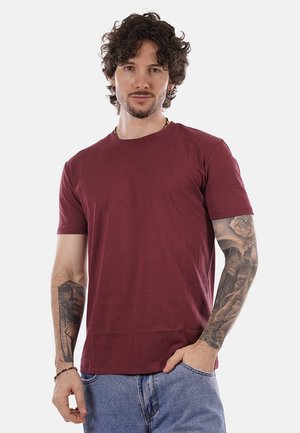 Uomo con capelli ricci che indossa una maglietta color prugna e jeans blu, che mostra tatuaggi dettagliati su entrambe le braccia, in piedi con una mano in tasca.