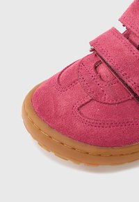 Ροζ παπούτσι από σουέντ με στρογγυλεμένη μύτη, που διαθέτει δύο λουράκια με βέλκρο και καφέ λαστιχένια σόλα. Ορατές ραφές και διακοσμητικά σχέδια.