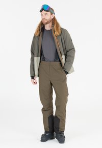 Mand med olivgrønt skijakke, mørkegrå skjorte, brune skipants med seler, sorte skistøvler og skibriller på panden.