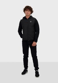 Chaqueta con capucha negra y cierre, con panel superior texturizado, combinada con pantalones negros y zapatillas negras con suelas blancas.