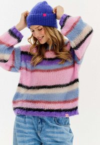 Žena upravuje fialovou pletenou čepici, má na sobě volný svetr s mnoha barvami a pruhy a světle modré džíny na jednobarevném pozadí.