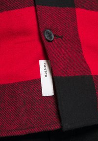 Camicia in flanella a quadri rossi e neri con chiusura a bottoni, tessuto texturizzato e etichetta di cura visibile sulla cucitura laterale.