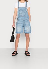 Ljusblå denimshorts med overalldesign har justerbara remmar, knappdetaljer och framfickor, som kombineras med en vit T-shirt och svarta sandaler.