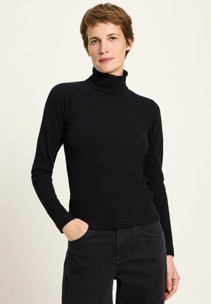 Frau mit kurzen braunen Haaren, die einen schwarzen gerippten Rollkragenpullover und schwarze Hosen trägt, steht mit einer Hand in der Tasche.