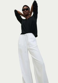 Czarna koszula z długim rękawem, białe spodnie w szerokim kroju z troczkami. Modelka w ciemnych prostokątnych okularach przeciwsłonecznych, z uniesionymi rękami, na neutralnym tle.