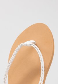 Valkoiset letitetyt hihnat flip-flopit, joissa on vaaleanruskea kumipohja. Littee muotoilu, jossa on sileä pinta ja pyöristetty varvasmuoto.