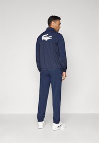 Bărbat îmbrăcat într-un trening bleumarin cu un logo alb de sneaker pe spatele jachetei, stând în fața unui fundal simplu de culoare gri deschis, cu corpul întors lateral.