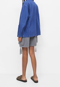 Niebieska kurtka dżinsowa z długimi rękawami, zestawiona z szarymi szortami typu cargo z bocznymi kieszeniami oraz wzorzystymi sandałami na płaskiej podeszwie.