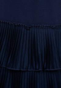 Granatowa plisowana tkanina o teksturowanej powierzchni, z falbaną u dołu i gładką górną częścią, prezentująca warstwowy design.
